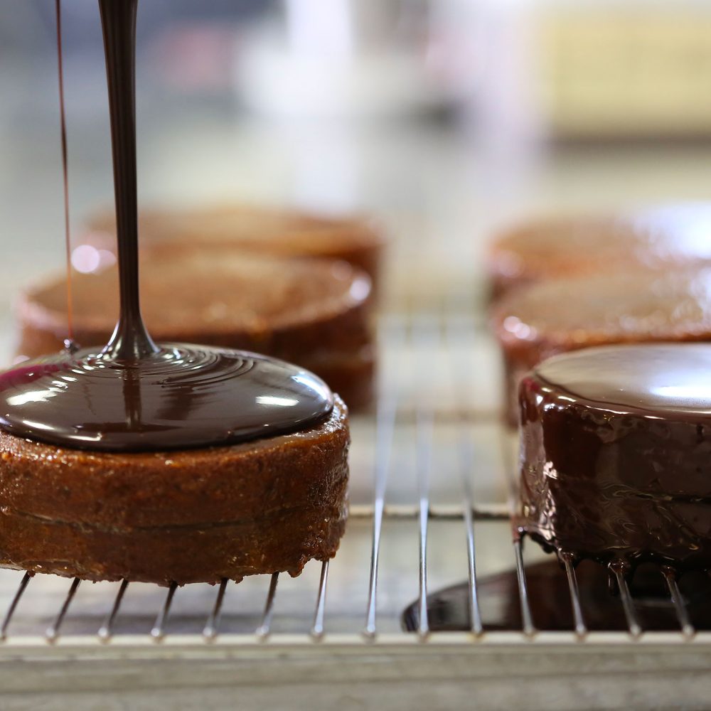 Fase di glassaggio artigianale con cioccolato fondente fuso su soffici tortine monoporzione, una lavorazione di precisione che garantisce una copertura lucida e perfetta, realizzata dalla Pasticceria Martinangeli (Le Dolci Tradizioni).