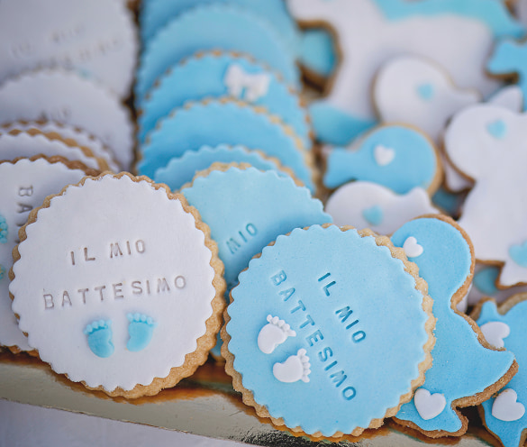 Assortimento di biscotti artigianali decorati con glassa bianca e azzurra per un battesimo, con la scritta "IL MIO BATTESIMO" e piccoli piedini decorativi, realizzati dalla Pasticceria Martinangeli (Le Dolci Tradizioni).