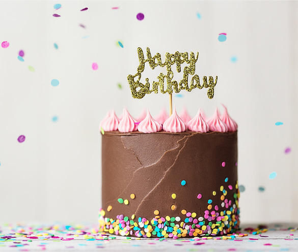 Torta di compleanno artigianale ricoperta di crema al cioccolato, decorata con ciuffetti di panna rosa, una pioggia di confettini colorati alla base e un topper dorato glitterato con la scritta "Happy Birthday", circondata da coriandoli colorati.