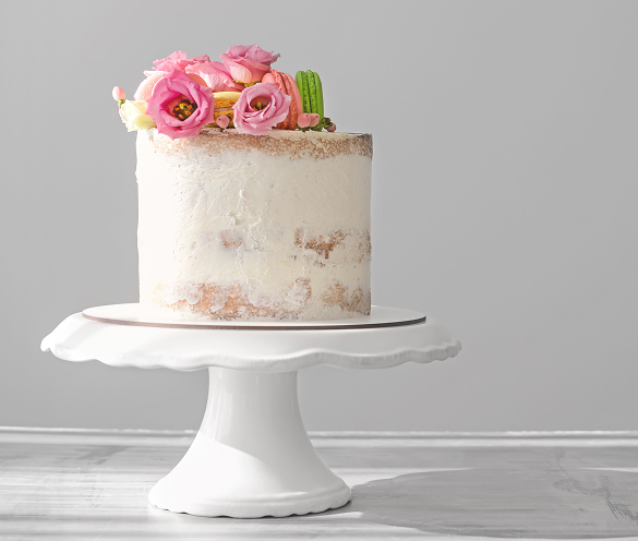 Elegante torta per comunioni artigianale con sottile copertura di panna bianca, decorata sulla sommità con fiori freschi nei toni del rosa e macaron colorati, presentata su un'alzata bianca.