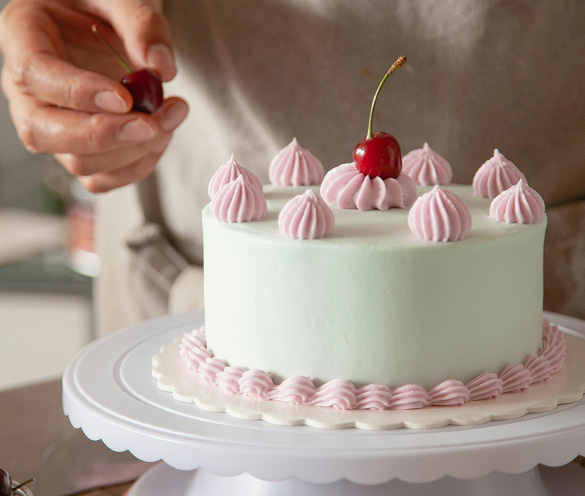 Torta artigianale con copertura verde menta, decorata con ciuffi di crema rosa e ciliegie fresche, mentre una mano ne aggiunge una sulla sommità.