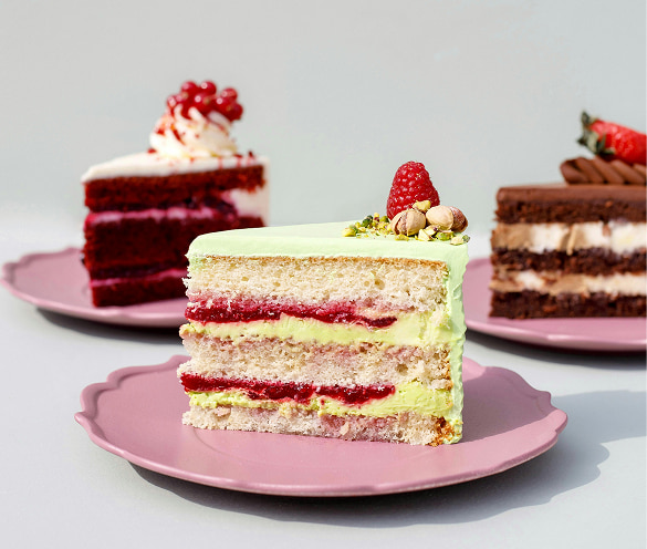 Selezione di fette di torta artigianali della Pasticceria Martinangeli (Le Dolci Tradizioni): in primo piano una fetta di torta a strati con crema al pistacchio e lamponi, accompagnata sullo sfondo da una fetta di Red Velvet e una al cioccolato, tutte servite su piattini rosa.