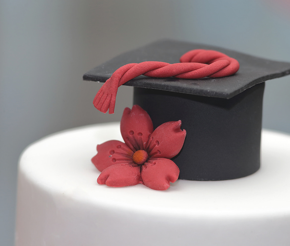 Dettaglio di una torta artigianale per laurea decorata con un tocco nero in pasta di zucchero, nappa rossa intrecciata e un fiore rosso alla base.