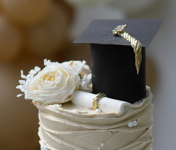 Dettaglio di una torta artigianale per laurea con frosting beige, decorata con un tocco nero in pasta di zucchero, nappa dorata, una grande rosa bianca e un piccolo diploma arrotolato.
